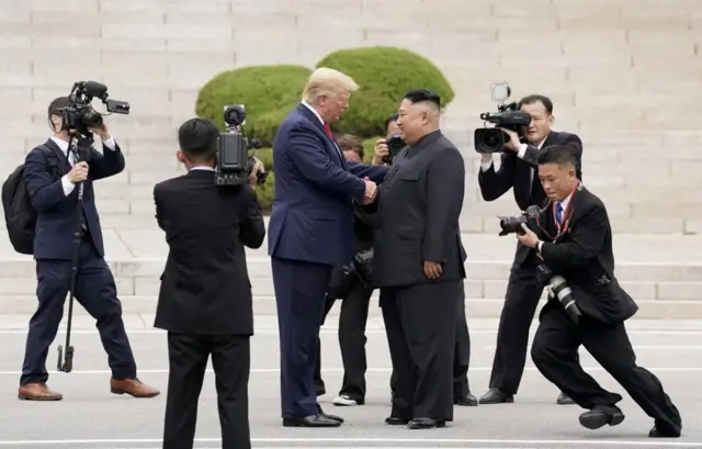 US President Donald Trump is seen shaking the hand of North Korea's leader Kim Jong Un. Trump is dressed in a navy suit with a red tie, Kim Jong Un is wearing a black suit. The two men are facing each other, they are surrounded by six photographers and camera men who are trying to capture the moment. The photographers are in motion, their dynamic poses indicate the importance of this moment. In the background there are white marble steps, and a blurred image of a small green bush. 
