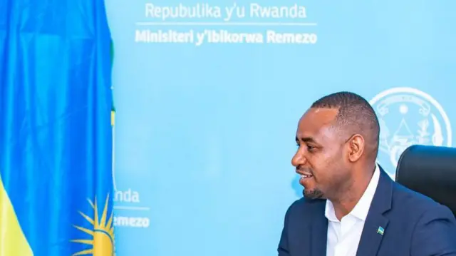 Umunyamabanga wa leta muri minisiteri y'ibikorwa-remezo Jean de Dieu Uwihanganye, yicaye yambaye ikositimu amwenyura, iruhande rwe hari ibendera ry'u Rwanda, hejuru ye hari ikirango cy'iyo minisiteri. 
