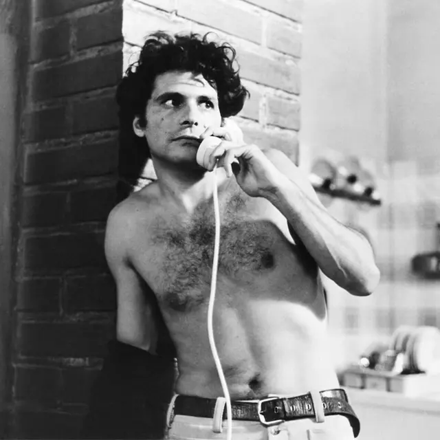 Un hombre sin camisa habla por telefono. Tiene cabello rizado, está despeinado y tiene el rostro serio. La imagen es en blanco y negro. 