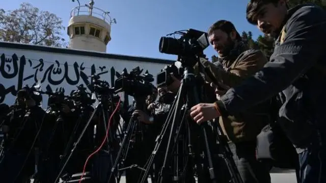 شماری از خبرنگاران در مقابل دیوارنوشته‌ای الله و اکبر. خبرنگاران در عقب کمره‌های شان در یک صف قرار دارند. 