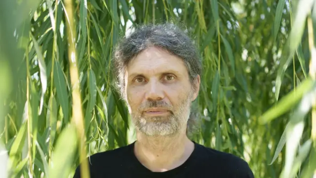 Мушкарац са дужом косом гледа у камеру, иза њега је зеленило, има седу косу и браду