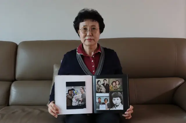 Han Tae-son sentada en un sofá, sosteniendo un álbum de fotografías abierto con fotos de su hija cuando era niña y cuando se reencontraron 44 años después de estar separadas.