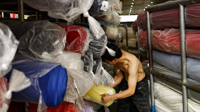 Guangzhou'daki bir tekstil deposunda, siyah pantolonlu ve üstsüz bir adam, plastik ambalajlara sarılı büyük kumaş rulolarıyla uğraşıyor.