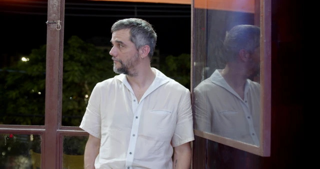 Wagner Moura está com camisa branca e se apoia em janela, com imagem refletida por espelho