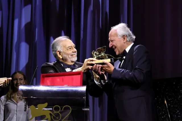 Coppola Herzog'a ödülü uzatıyor, ikisi de smokin giymiş, gülümsüyorlar.