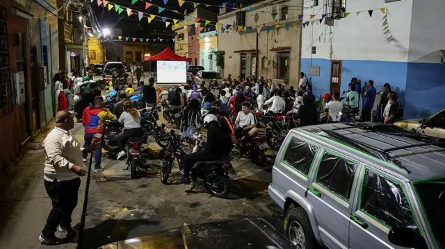 Un grupo de personas están reunidas en medio de una calle de un barrio humilde de Caracas, mirando una pantalla.