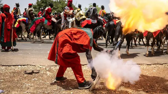 Smoke and a bright orange light can be seen as a man, wearing red and green, fires a gun