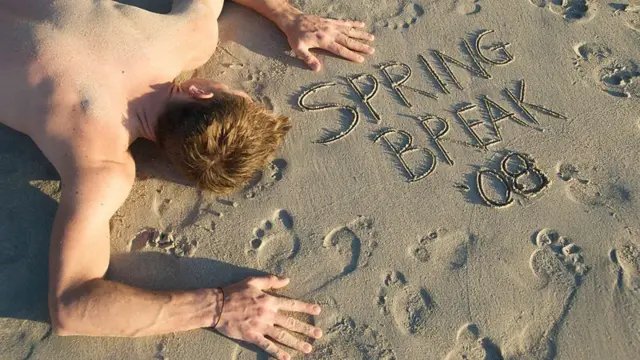 Homem sem camisa deitado de bruços na areia de uma praia parecendo estar desmaiado após ter bebido demais, ao lado dele está escrito na areia 'Férias de Primavera 2008' em inglês