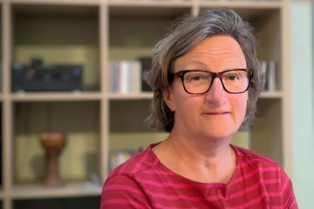 Sarah Hendrickx, que está sorrindo, usando óculos e em frente a um conjunto de prateleiras