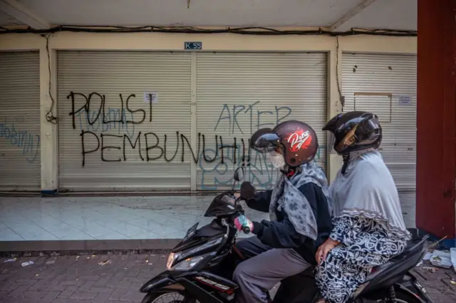 Para wanita mengendarai sepeda motor mereka melewati grafiti bertuliskan 'polisi pembunuh' di stadion Kanjuruhan, lokasi penyerbuan mematikan, di Malang, Jawa Timur pada 4 Oktober 2022. - 