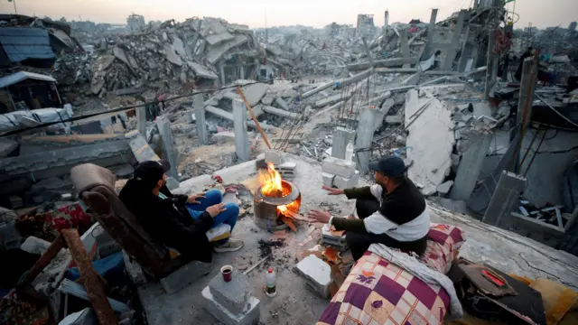 فلسطينيون يجلسون بجوار النار بين أنقاض المباني التي دمرت خلال الهجوم الإسرائيلي، بعد وقف إطلاق النار بين إسرائيل وحماس، في مخيم جباليا للاجئين، شمال قطاع غزة، 17 فبراير/شباط 2025.