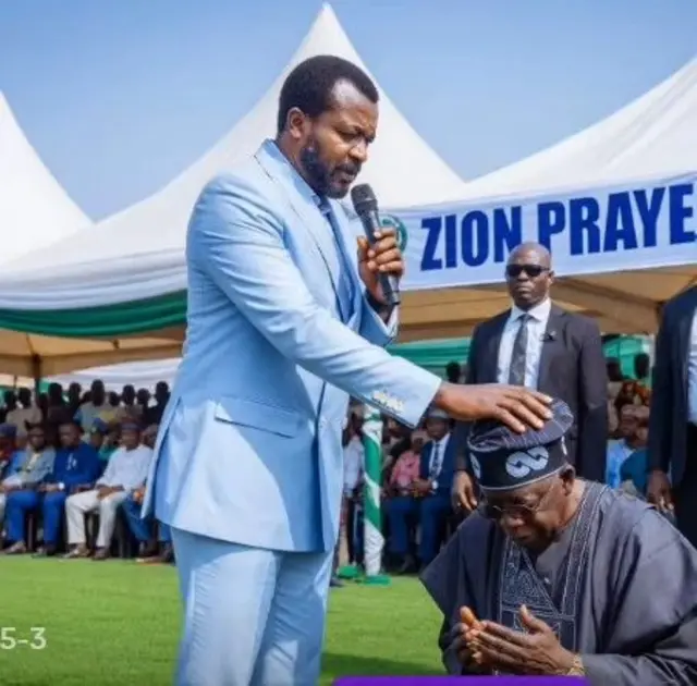 Pasitọ to wọ aṣọ oyinbo gbee ọwọ le Aarẹ Tinubu lori ninu aworan yii, nigba to fi han pe Aarẹ kunlẹ pẹlu.