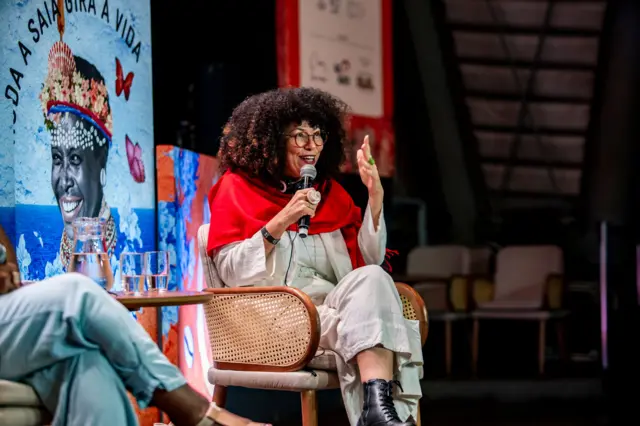 Aparecida gesticulando e sorrindo enquanto fala em evento, sentada