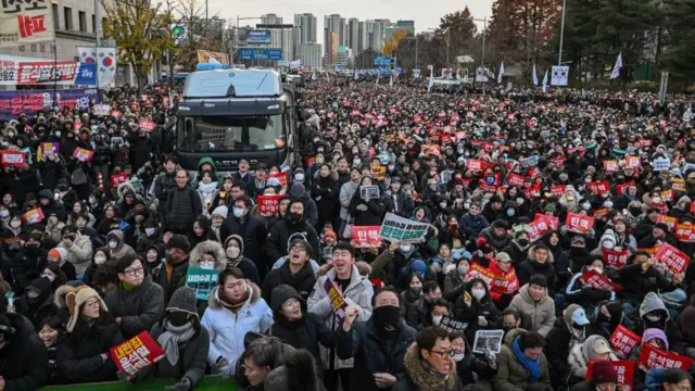 2024年12月7日，人们在首尔国会外参加示威活动，要求罢免韩国总统尹锡悦。大量身着冬装的人群手持海报，挥舞着韩国国旗。中间是一辆黑色货车，可能是警车。