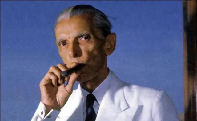 সাদা কোট পড়া সাদা চুলের জিন্নাহর হাতে একটি সিগার যেটি তিনি ঠোঁটের কাছে ধরে রেখেছেন