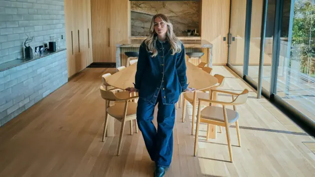 Mickey Ross denim bir gömlek ve kotla masaya yaslanmış. 