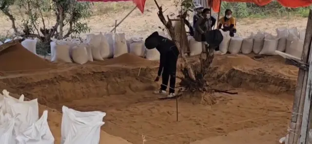 පුද්ගලයෙකු කැණීම් සිදුකෙරන ස්ථානයේ සිටගෙන සිටින අතර අනෙක් පසින් ඉවත් කරන ලද පස් අඩංගු පොලිතීන් උර කිහිපයක් දකින්නට ලැබේ.
