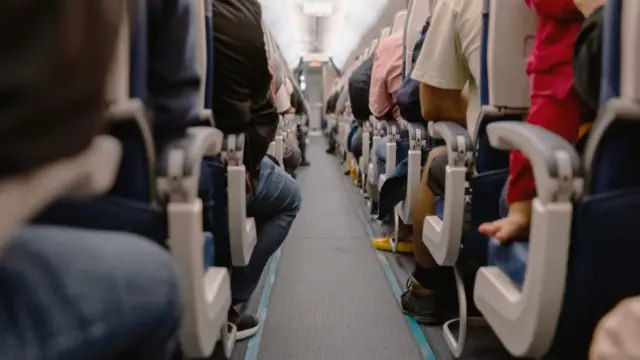 Corredor em meio a duas fileiras de assentos de um avião com muitos passageiros a bordo