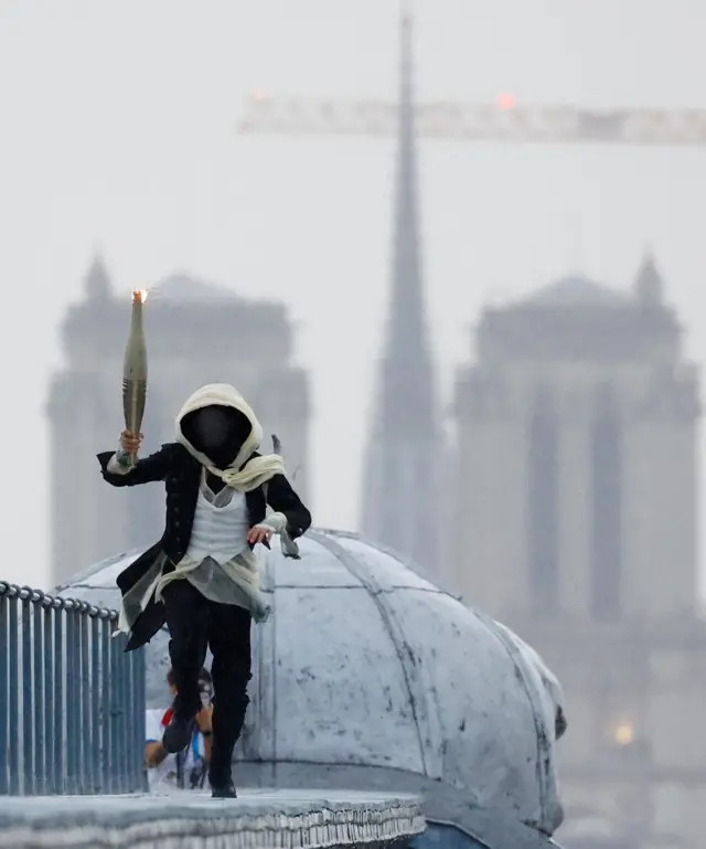 Un porteur de torche masqué court au sommet du musée d'Orsay avec la cathédrale Notre-Dame-de-Paris en arrière-plan lors de la cérémonie d'ouverture des Jeux olympiques Paris 2024, le 26 juillet 2024.