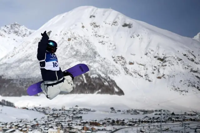 上半身が黒っぽい色、下半身が白のウエアを着て、ヘルメットとゴーグルをつけた長谷川が、空中で右手を上げている。背後には雪景色の山と集落が見える