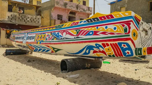 One colourful fishing canoe from di back angle. E dey sit on rollers on di beach. We fit see some buildings for di background of di beachside. 