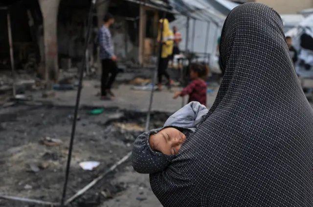 A Palestinian woman carries a child, while Palestinians inspect the damage to a school that houses the displaced, after an Israeli raid, in Gaza City, April 23, 2025.