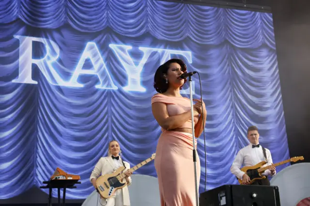 Raye stands on stage singing, in a pink dress, in front of a backdrop that says her name in capital letters.