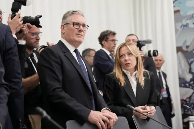 Starmer ve Meloni sandalyelerin arkasında ayakta durmuş konuşuyorlar. Arkalarında da insanlar ve gazeteciler sıralanmış. 