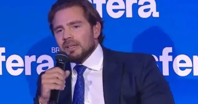 Foto de Daniel Vorcaro usando terno azul marinho, camisa branca, gravata azul, segurando um microfone. É um homem branco de barba e cabelos escuros.