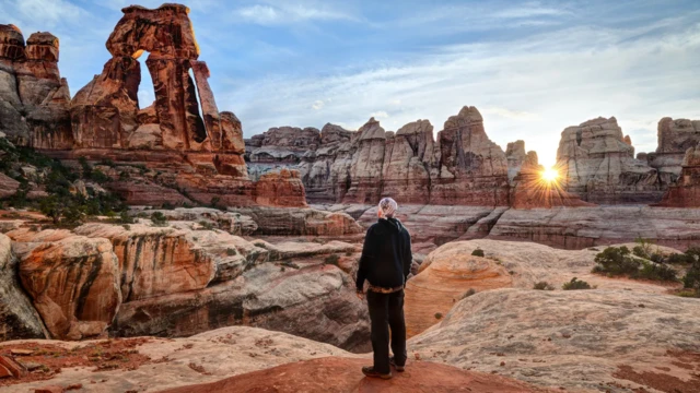 Park narodowy Canyonlands o wschodzie słońca. Odwrócony plecami do aparatu turysta podziwia żółtobrązowe formacje skalne. W tle niebieskie, lekko zachmurzone niebo i widoczne nisko na horyzoncie słońce