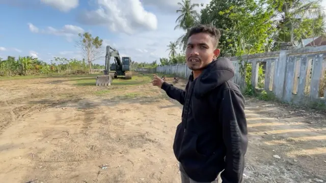 Faizil menggunaka hoodie berwarna hitam dan jeans abu-abu, berdiri di lahan kosong yang cukup luas sambil menunjuk alat berat yang ada di sekitarnya.