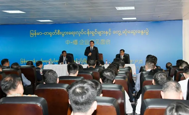 လုပ်ငန်းရှင်တွေလို့ ယူဆရတဲ့ အမျိုးသား ၂၀ နီးပါး ကင်မရာကို ကျောပေးပြီး ထိုင်ခုံတွေမှာ ထိုင်နေ၊ စင်မြင့်ထက်မှာတော့ အနောက်တိုင်း ဝတ်စုံဝတ်ထားတဲ့ ဦးညိုစောက မိုက်ကရိုဖုန်းကိုင်ပြီး မတ်တပ်ရပ်နေ၊ သူနဲ့အတူ စင်မြင့်ထက်မှာ အမျိုးသား ၂ ဦးလည်း တစ်ဖက်တစ်ချက်စီ ထိုင်နေကြ။ သူတို့အနောက်က နံရံမှာတော့ မြန်မာ -တရုတ် စီးပွားရေး လုပ်ငန်းရှင်များ တွေ့ဆုံဆွေးနွေးပွဲဆိုတဲ့ အပြာရောင်အောက်ခံပေါ် အဝါရောင် စာသားကို တရုတ် မြန်မာ နှစ်ဘာသာနဲ့ရေးထားတာတွေ့ရ၊ နန်နင်းမြို့ဆိုတဲ့စာသားနဲ့ ၂၀၂၅ ခုနှစ် ၉ လပိုင်းဆိုတဲ့ စာသားကိုလည်း တစ်စိတ်တစ်ပိုင်းမြင်ရ 