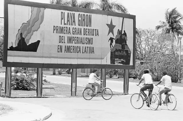 Tres personas en bicicleta pasan junto a un cartel de propaganda que exalta la 