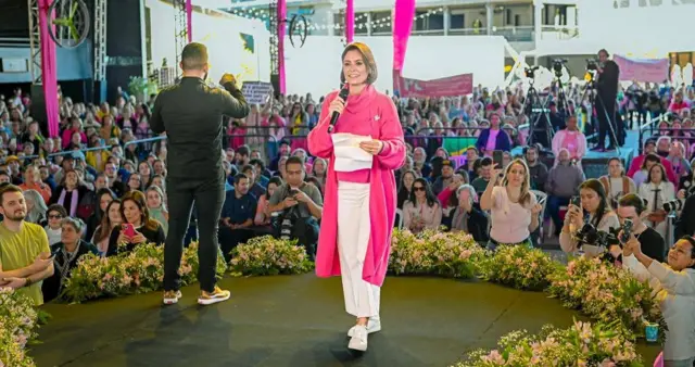 Encontro do PL Mulher, com a presença de Michelle Bolsonaro, em Santa Catarina, em 29 de julho de 2023. No palco cercado de flores rosas, Michelle veste rosa e fala ao microfone, ao lado de um tradutor de libras vestido de preto, com a plateia ao fundo