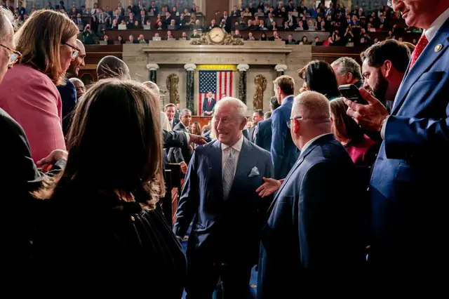 У великій парламентській залі людина в темному формальному костюмі стоїть у проході серед скупчення присутніх. Навколо — люди в діловому одязі, частина з них фотографує або знімає на телефони. На задньому плані видно трибуну, великий прапор США та заповнені глядацькі ряди; атмосфера офіційна й багатолюдна.