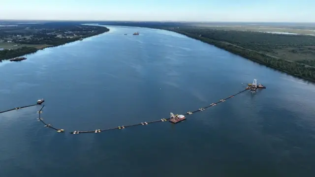 Plan large d'une rivière qui s'étire au loin, bordée d'arbres, de champs et de bâtiments ; au premier plan, plusieurs embarcations naviguent, reliées par une longue ligne qui s'étend sur presque toute la largeur de la rivière.