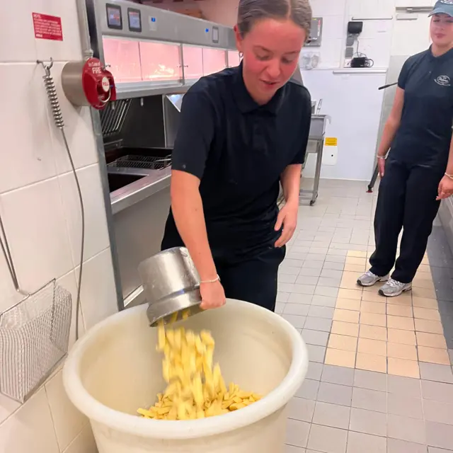 Kenzie vierte las patatas fritas de un recipiente de acero inoxidable en un cubo grande de plástico blanco en la freidora. Las freidoras y las vitrinas de comida caliente están detrás de ella, de cara a la zona de clientes.
