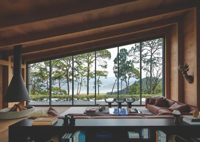 Sala de estar con estructura de madera, chimenea suspendida y ventanales panorámicos con vista a un lago entre pinos.