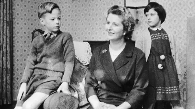 Тэтчер сидит на кресле, слева от нее на ручке кресла сидит ее сын, справа дочь. На заднем плане виден интерьер квартиры, камин и занавески на окне.