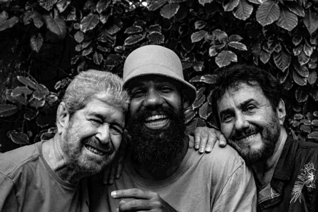 Dupla sorrindo para a foto com Russo Passapusso; eles estão próximos, abraçados, em frente a muro com plantas
