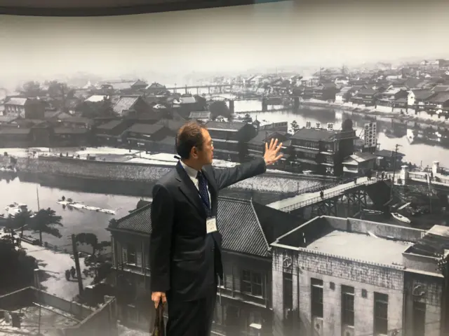 Мушкарац руком показује на фотографију Хирошиме пре бомбардовања