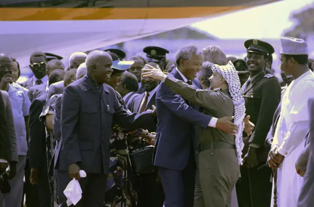 Mwenyekiti wa PLO Yasser Arafat akimkumbatia Rais wa Afrika Kusini Nelson Mandela, tarehe 1 Januari 1990, huku wangine wakitabasamu. 