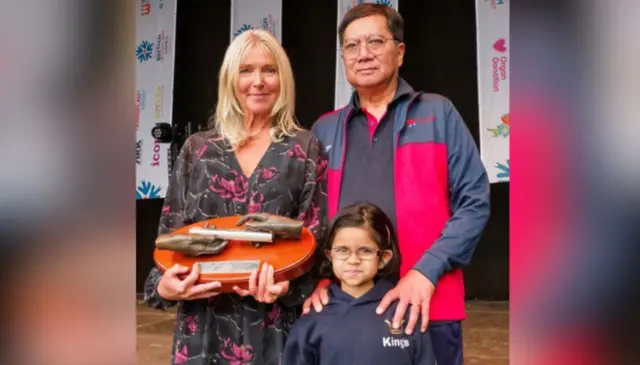 ليزا ويلسون في صورة مع المتلقين للأعضاء غوردون وفاطمة. السيدة ويلسون تحمل عصا توم، شعرها أشقر وترتدي فستاناً أسود عليه ورود كبيرة بلون وردي غامق. إلى يمين الصورة بجانبها تماماً يقف غوردون ذو الشعر الأسود، مرتدياً نظارات طبية ولباساً رياضياً يبدو أن اللون الغالب عليه هو الكحلي وفيه بعض الأجزاء لونها بين الأحمر والوردي. ويمسك بكتفي فاطمة التي تقف أمامه ترتدي نظارات طبية ولباساً رياضياً يبدو كحلي اللون، وشعرها أسود.