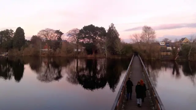 Una pasarela de madera con tres personas cruzando. Parte del cielo es morado, lo que se refleja en el agua.