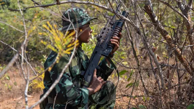 Seorang pemuda berseragam tempur berlutut dengan senapan serbu di semak-semak