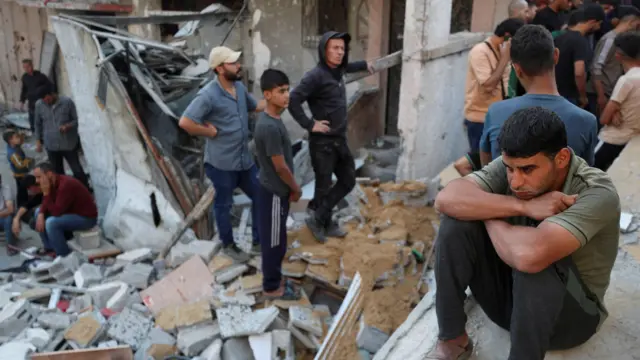 Um homem sentado enquanto palestinos se reúnem no local de um ataque israelense a uma casa em Gaza.