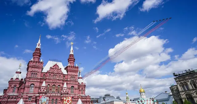 Над Червоною площею в ясному небі летить група літаків, залишаючи за собою смуги у кольорах російського прапора. Внизу видно червоні цегляні будівлі з вежами, поруч — золоті куполи храму.