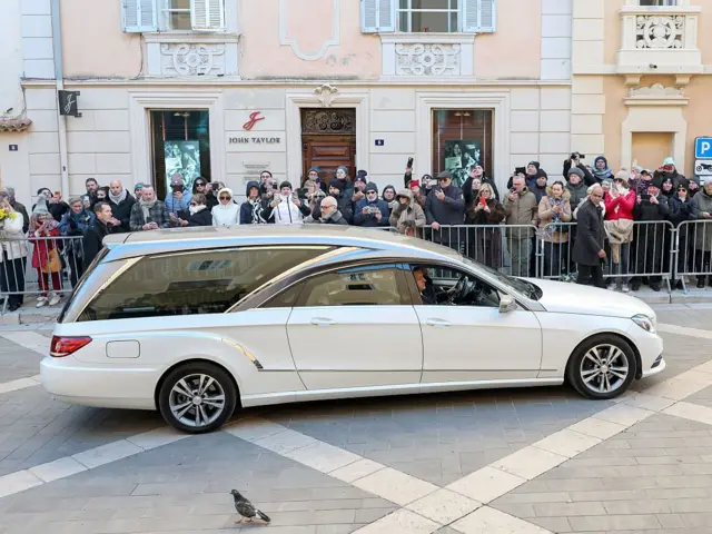 Катафалк с гробом едет к церкви, за ограждением наблюдают люди.