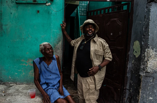 El líder pandillero Jimmy Chérizier, conocido como Barbecue, líder de Viv Ansanm, vestido con camisa, pantalón y gorra beige junto a una anciana que viste de azul en un barrio de Puerto Príncipe, Haití, el 6 de julio de 2026.
