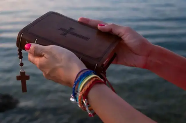Las manos de una mujer con manillas de los colores de la bandera gay cargan una biblia, de la cual cuelga un rosario.